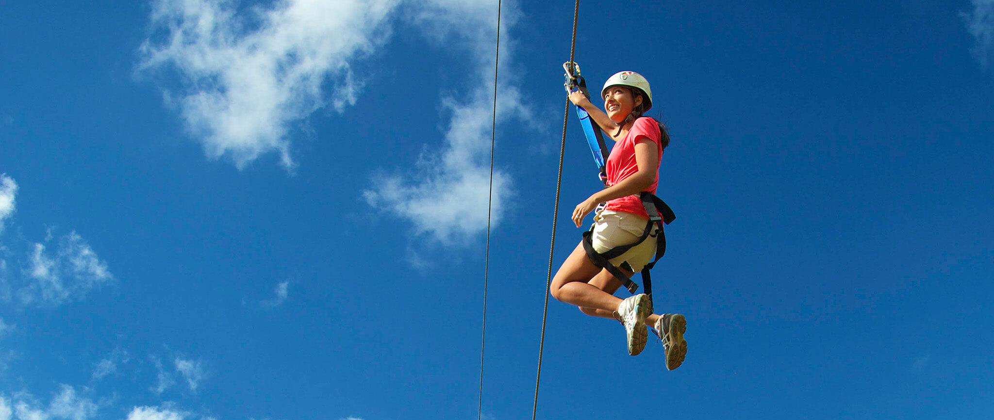 Woman ziplining with blue sky and white clouds in the background.