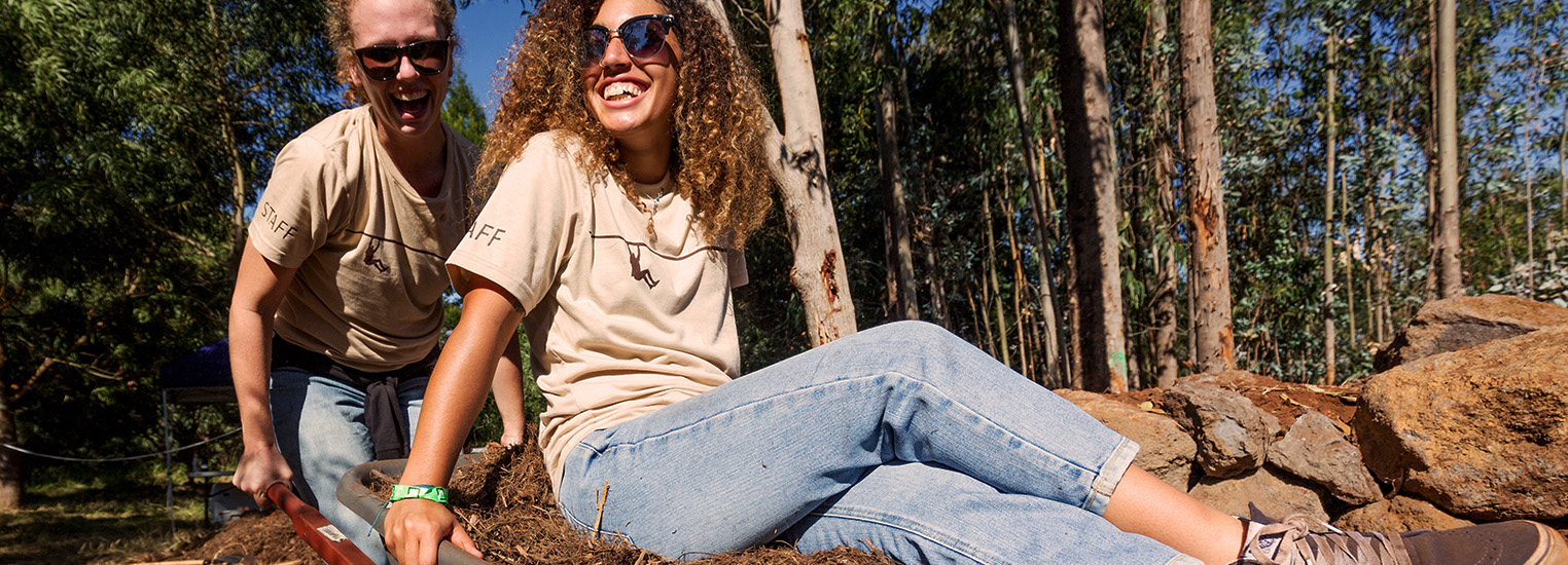 Two women laughing. The first is hauling a wheelbarrow filled with dirt and park. The second women is also sitting in the wheelbarrow